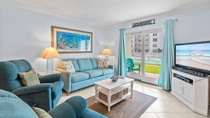 A living room with a sofa, a coffee table, a TV, and a painting. - Sea Coast Gardens II 110 (New Smyrna Beach)
