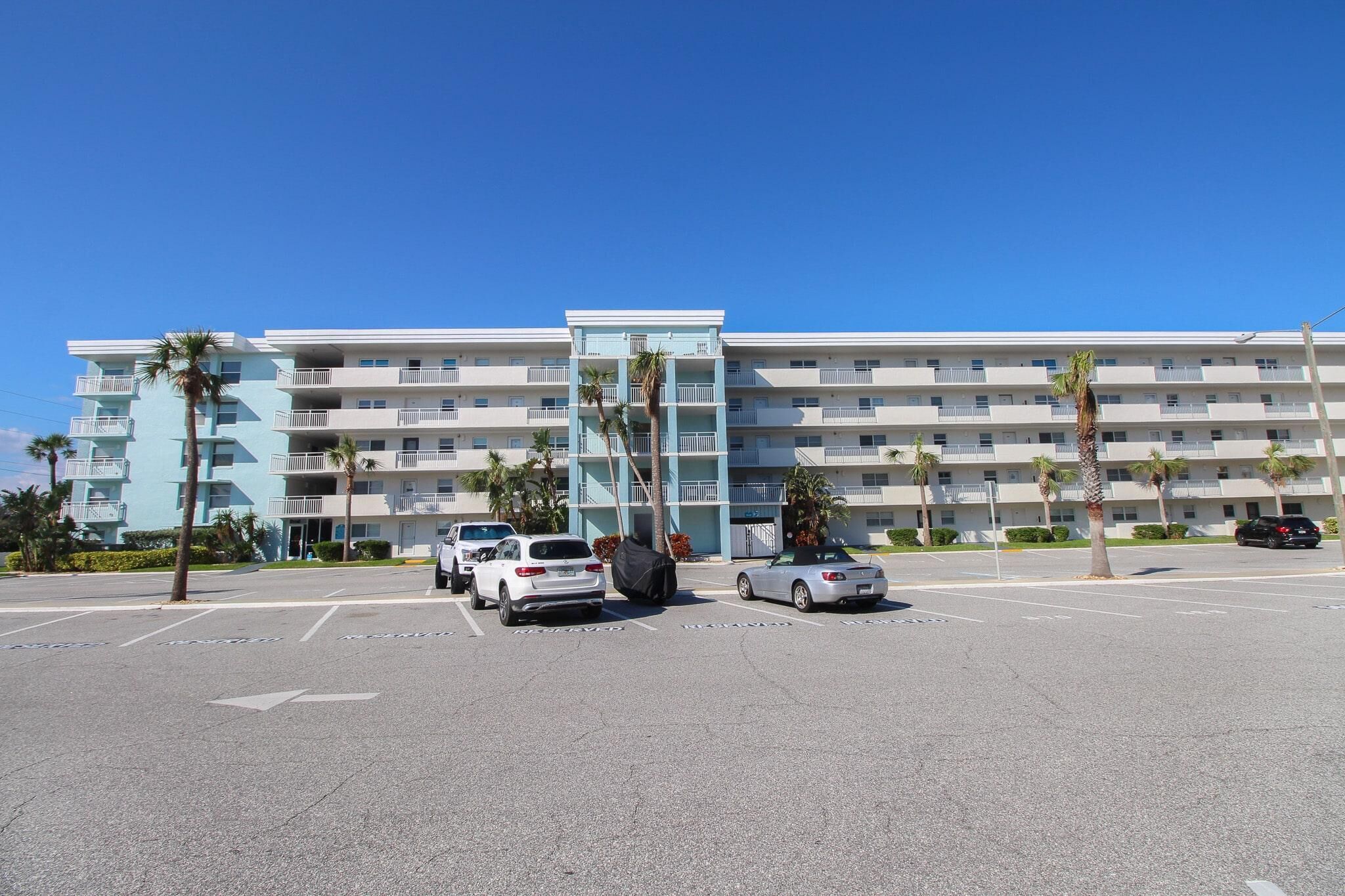 A multi-story hotel with balconies, a parking lot, and palm trees.