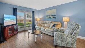 A living room with a TV, leather sofa, patterned armchair, and a view of the beach through a sliding door. - Sea Coast Gardens II 117 (New Smyrna Beach)