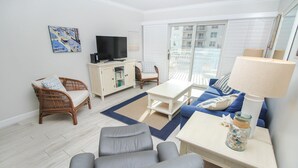 Una sala de estar moderna con un sofá gris, un sillón de mimbre, una mesa de centro blanca, un televisor sobre un mueble bajo y un balcón con vista a edificios.