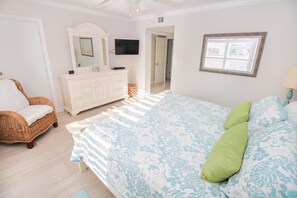 A bedroom with a bed, a dresser, a wicker chair, a mirror, and a television. - Sea Coast Gardens II 211 (New Smyrna Beach)