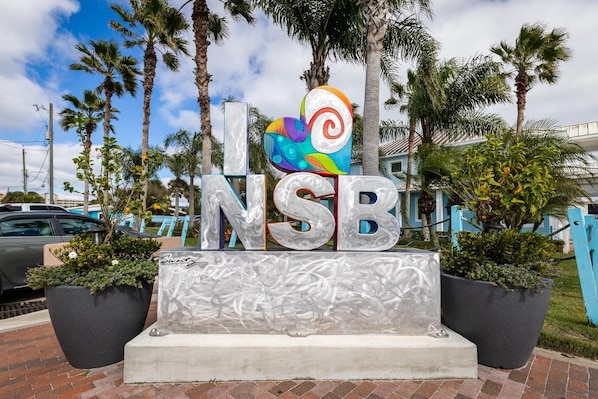 A sign with the letters "NSB" and a colorful design above it, surrounded by palm trees and a brick pathway. - Sea Coast Gardens II 211 (New Smyrna Beach)