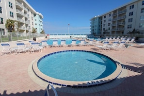 A circular pool with lounge chairs around it, a flag, and a view of a building in the background. - Sea Coast Gardens II 211 (New Smyrna Beach)