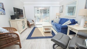 Una sala de estar con un sofá azul, un televisor sobre un mueble, sillas de mimbre, una mesita y vistas a través de una puerta de cristal.