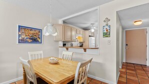 A dining area with a wooden table and white chairs, a pendant light, and a kitchen in the background. - Sea Coast Gardens II 307 (New Smyrna Beach)
