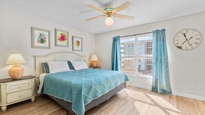 A bedroom with a bed, nightstands, a ceiling fan, a window with curtains, and a wall clock. - Sea Coast Gardens II 307 (New Smyrna Beach)