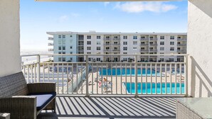 A balcony with a wicker chair, a pool area with lounge chairs, and a view of a hotel building. - Sea Coast Gardens II 307 (New Smyrna Beach)