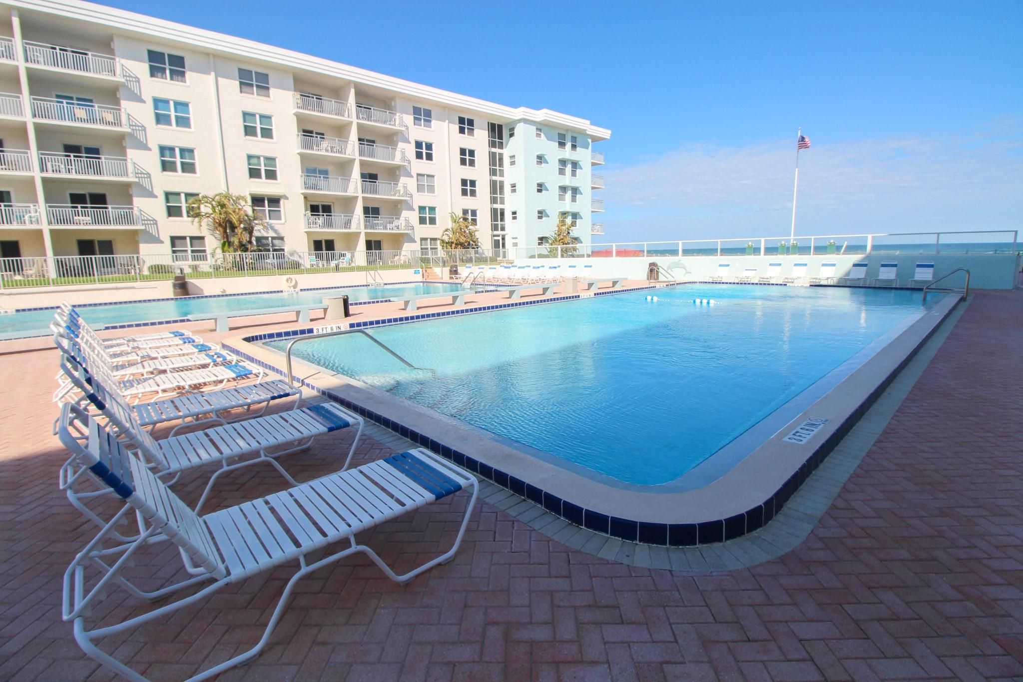 A swimming pool with lounge chairs, a tiled deck, and a multi-story building with balconies.