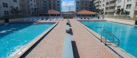 A hotel pool area with two swimming pools, lounge chairs, and a brick walkway.