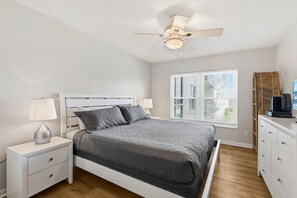 A bedroom with a bed, nightstands, a ceiling fan, a window with blinds, and a dresser. - Sea Coast Gardens II 316 (New Smyrna Beach)