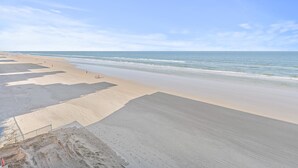 A wide sandy beach with gentle waves, a clear sky, and a few people in the distance. - Sea Coast Gardens II 402 (New Smyrna Beach)