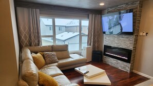 A modern living room with a leather sofa, a flat-screen TV mounted on a stone wall, and a fireplace. - beautiful, cozy private and quiet house + heat garage -EV charger - NO BASEMENT (Edmonton)