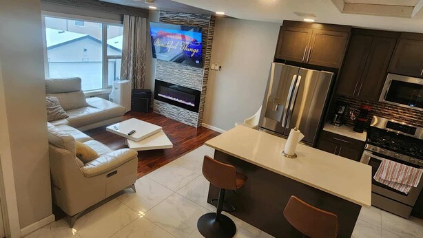 A modern living room with a sofa, a coffee table, and a TV. There is a kitchen area with a refrigerator, microwave, and stove. - beautiful, cozy private and quiet house + heat garage -EV charger - NO BASEMENT (Edmonton)