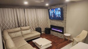 A modern living room with a beige leather sofa, a flat-screen TV mounted on a textured wall, a fireplace, and a small white table. - beautiful, cozy private and quiet house + heat garage -EV charger - NO BASEMENT (Edmonton)