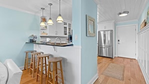Una cocina con isla central, taburetes de madera, iluminación colgante y un refrigerador de acero inoxidable.