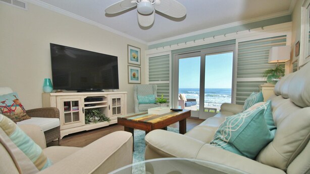 A living room with a television, a coffee table, and a sofa. There is a view of the ocean through large glass doors.