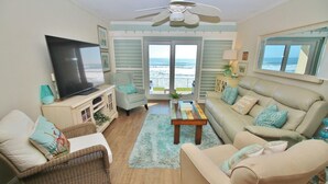 A living room with a television, wicker furniture, a beige sofa, and a view of the ocean through the windows.
