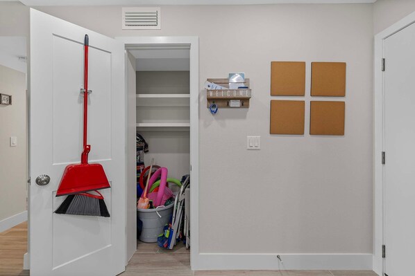 Un pasillo con una puerta cerrada, una escoba apoyada en la pared y un estante de almacenamiento con un letrero que dice 'WELCOME'.