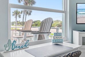 A beach-themed interior with a view of the ocean, two Adirondack chairs, a table, and a sign with 'Live Laugh Love' on a windowsill. - Sea Coast Gardens III 215 (New Smyrna Beach)