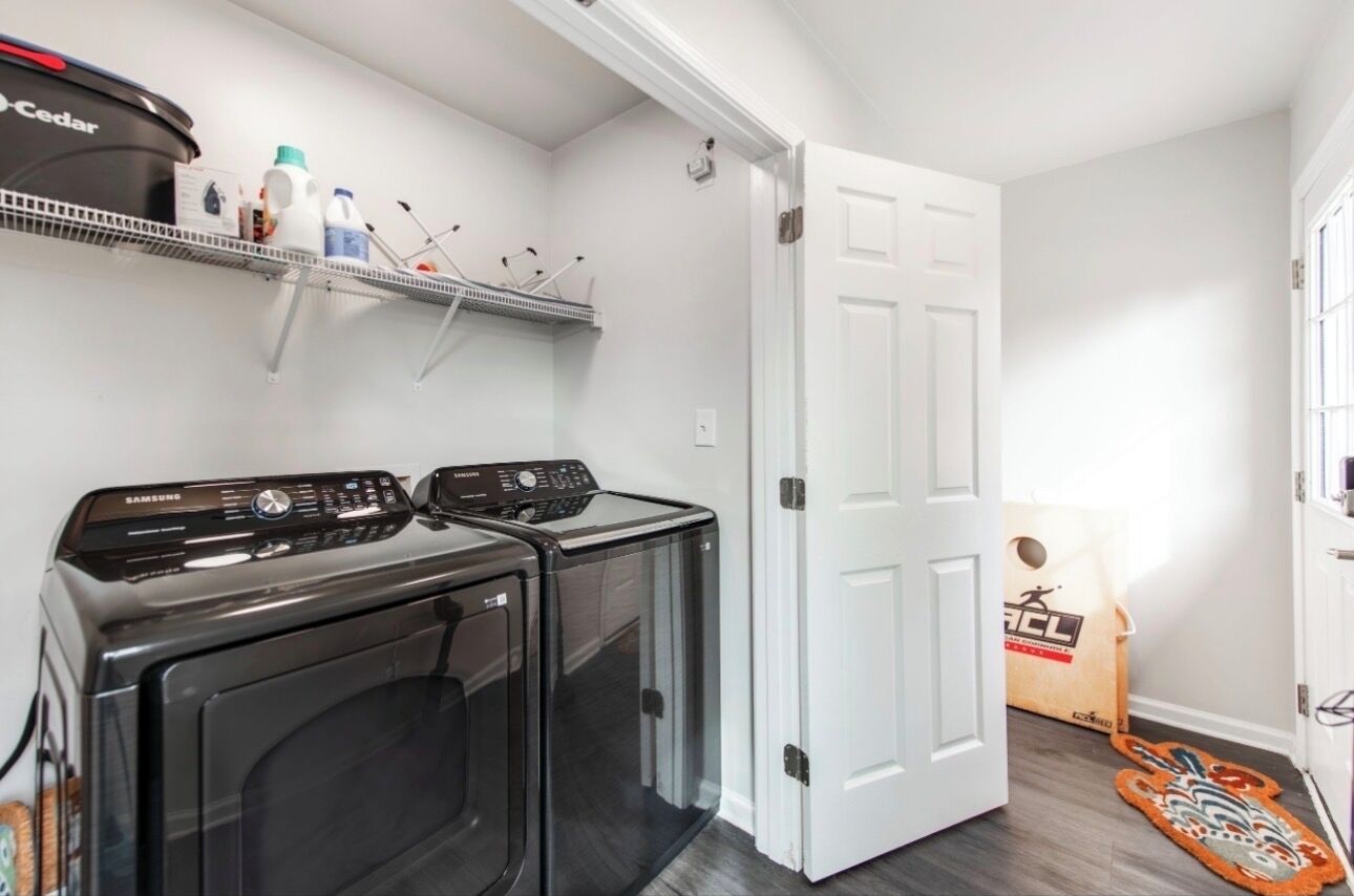 A laundry room with two Samsung washing machines, a shelf with cleaning supplies, and a door leading to another room.