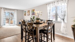 A dining area with a round table, six chairs, and a vase of flowers. There are two windows with curtains, a door leading outside, and artwork on the walls. - Welcome to Valley View (Derby)