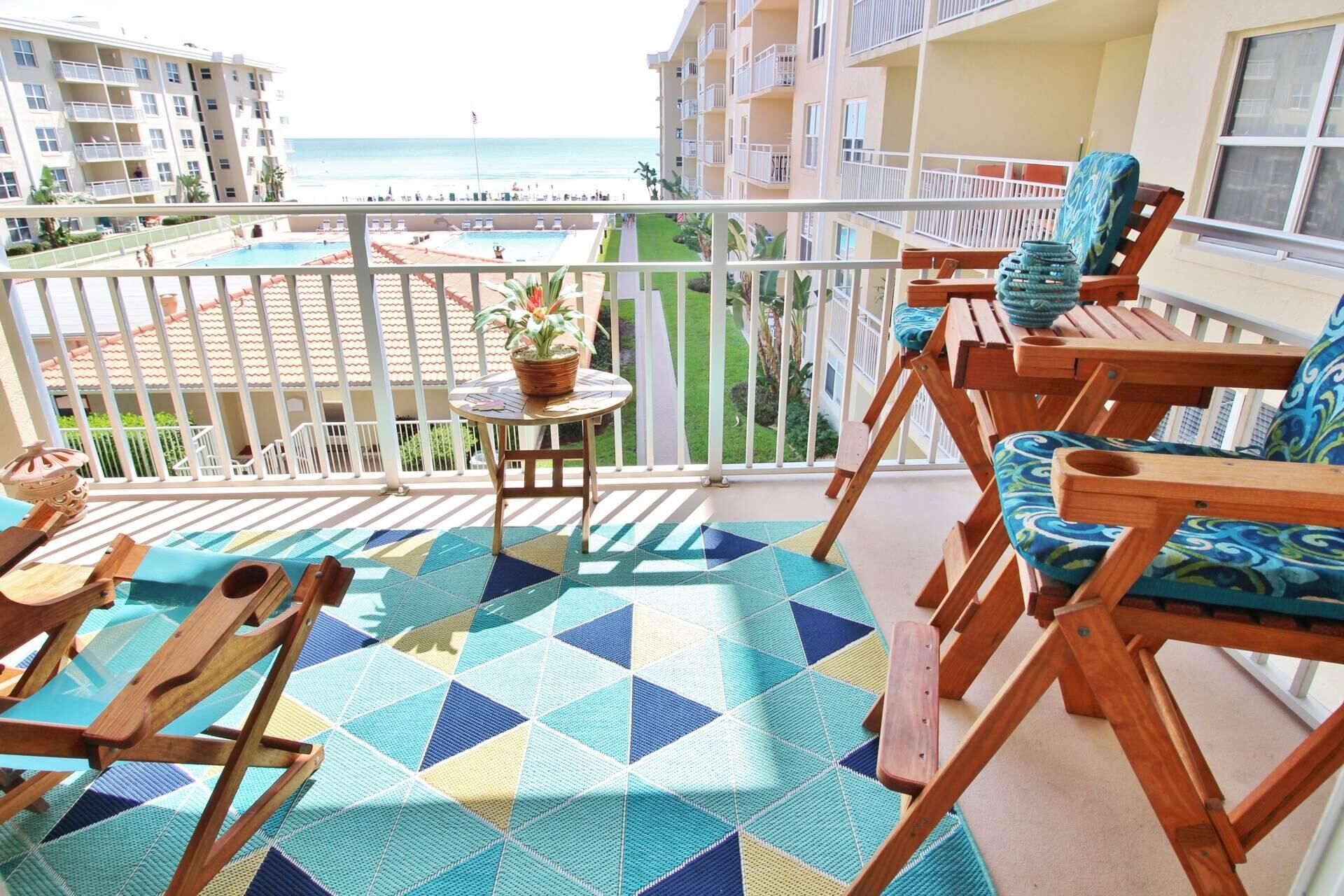 A balcony with a view of the beach, wooden chairs, a small table with a plant, and a patterned rug.