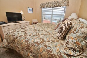A hotel room with a bed, a television on a dresser, a window with blinds, and a framed picture on the wall.