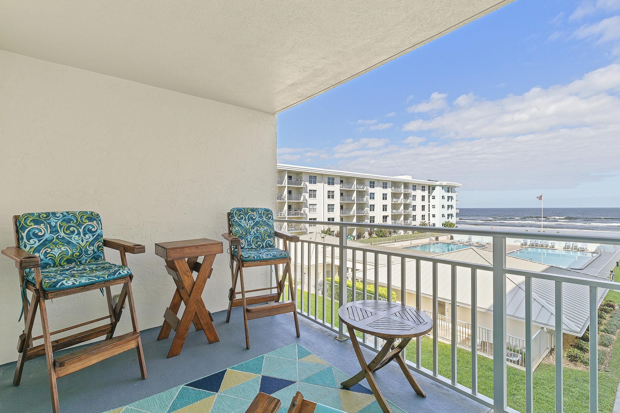A balcony with two wooden chairs, a small table, and a view of a building and beach.