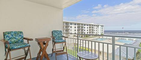 A balcony with two wooden chairs, a small table, and a view of a building and beach.