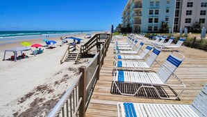 A beachside promenade with sun loungers and umbrellas, a wooden walkway, and a building in the background.