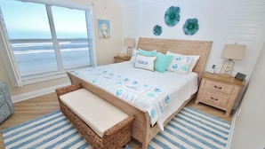 A bedroom with a large window overlooking the ocean, a bed with a beach-themed bedspread, a wicker bench, and a nightstand with a lamp and a clock. - Sea Coast Gardens III 406 (New Smyrna Beach)