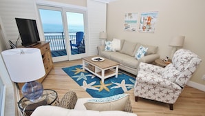 A living room with a sofa, armchair, and a coffee table. There is a TV on a stand, a lamp, and a chair with a view of the ocean. - Sea Coast Gardens III 406 (New Smyrna Beach)