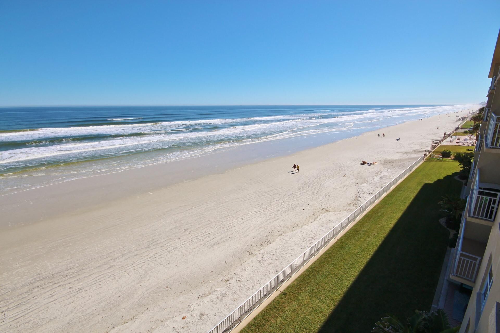 A beach with a clear view of the ocean, a few people, and a building with a balcony overlooking the beach.