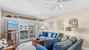 A living room with a view of the beach, a ceiling fan, and a sign that says 'BEACH HOUSE'.