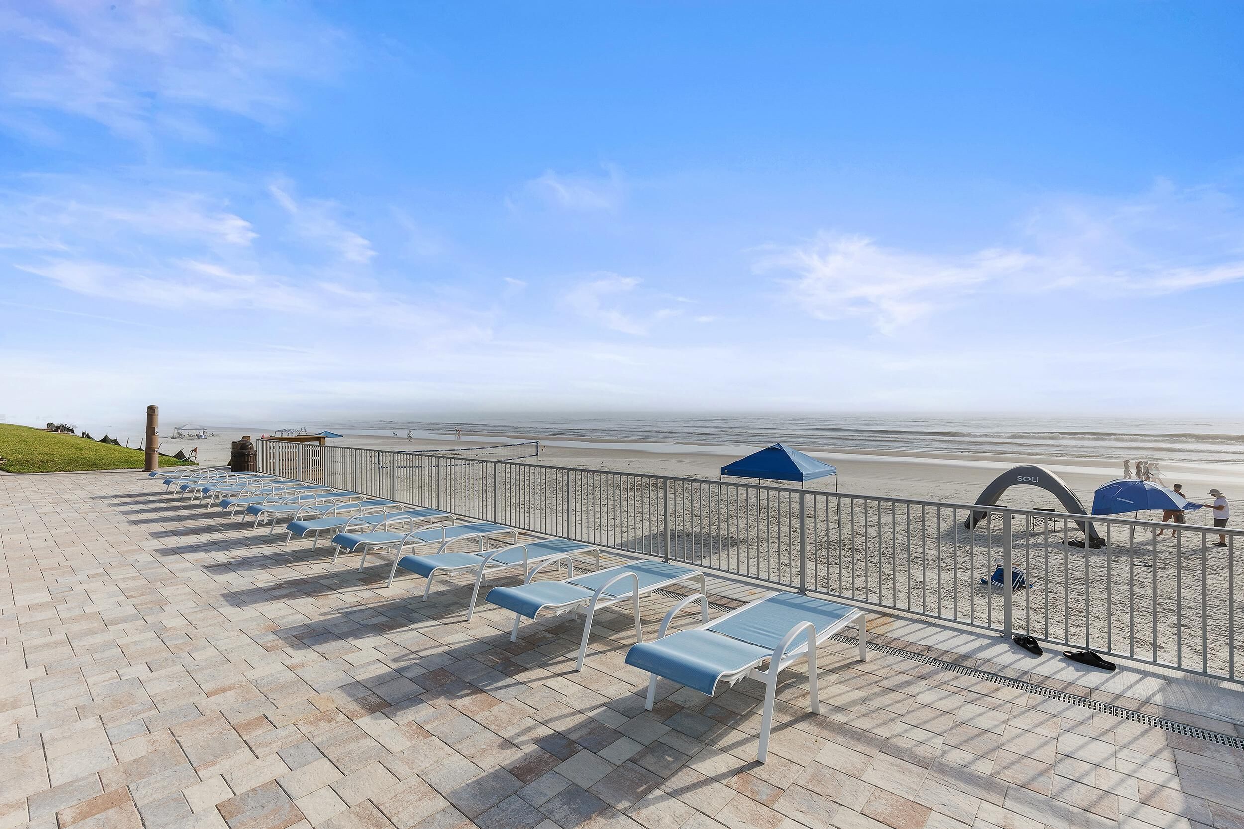 En strandnära lounge med solstolar och parasoller, ett metallräcke och utsikt över stranden och havet.