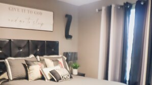 Un dormitorio moderno con una cama grande, cabecera, mesita de noche, lámpara de pared, ventana con cortinas y una frase inspiradora en la pared.