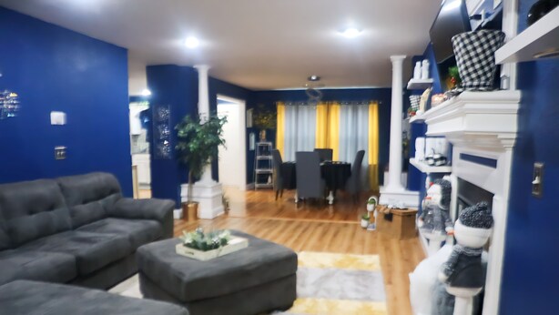 Una sala de estar con un sofá gris, un pequeño taburete y un tapete amarillo. Se observa un comedor con una mesa y sillas al fondo.