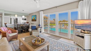 A spacious living room with a large TV, comfortable seating, a dining area, and a balcony with a pool view. - Schooner Point A204 (New Smyrna Beach)