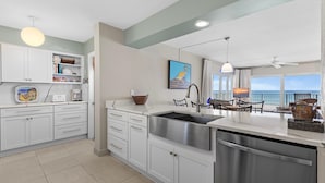 A modern kitchen with white cabinets, a stainless steel sink, and a dining area with a view of the ocean. - Schooner Point A204 (New Smyrna Beach)