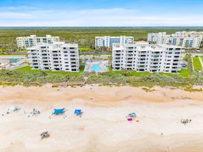 A beachfront hotel with multiple buildings, a swimming pool, and a sandy beach with umbrellas and beach chairs.