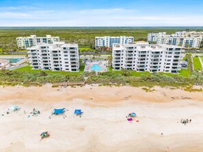 A beachfront hotel with multiple buildings, a swimming pool, and a sandy beach with umbrellas and beach chairs.
