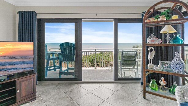 A living room with a TV, a bookshelf, and a view of the ocean through a sliding glass door.