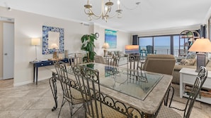 A spacious living area with a glass-top dining table, metal chairs, a chandelier, and a large window offering a view of the ocean.