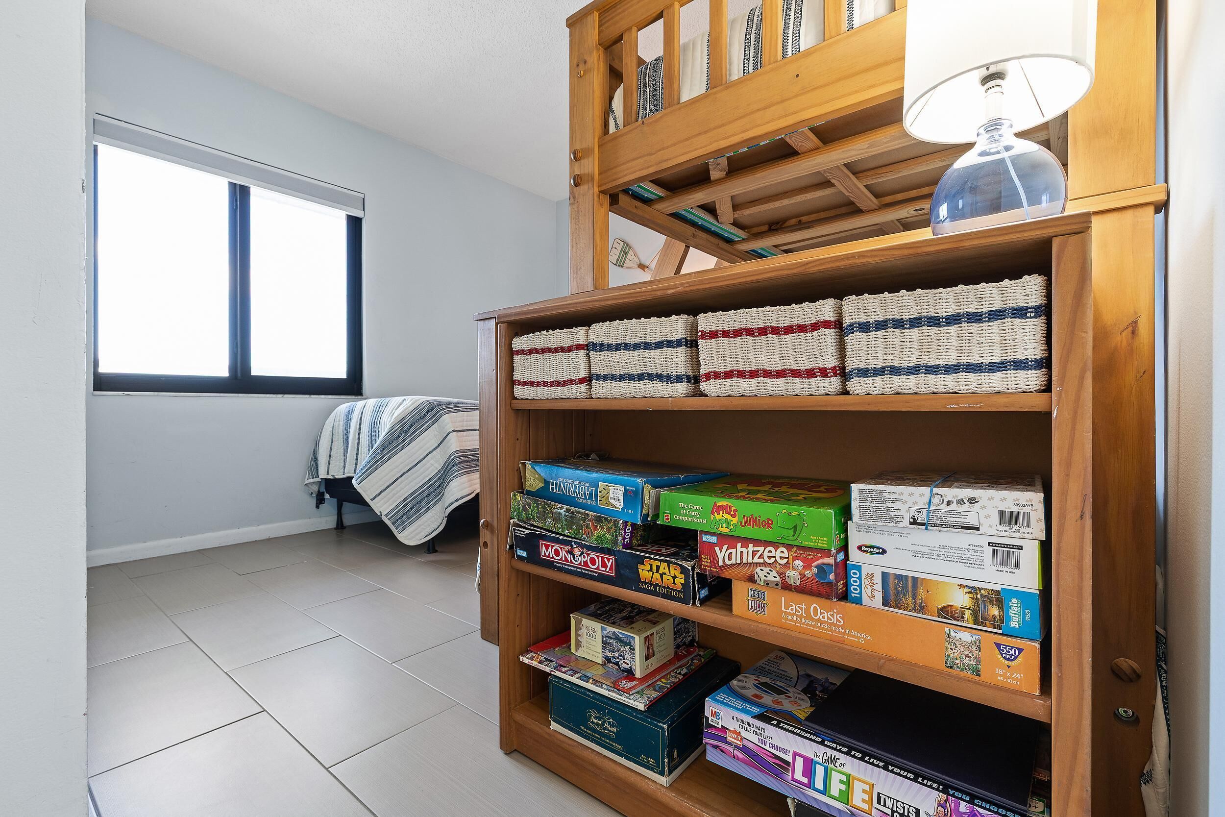 A room with a wooden bookshelf filled with board games, a window with curtains, and a bed with a striped blanket.