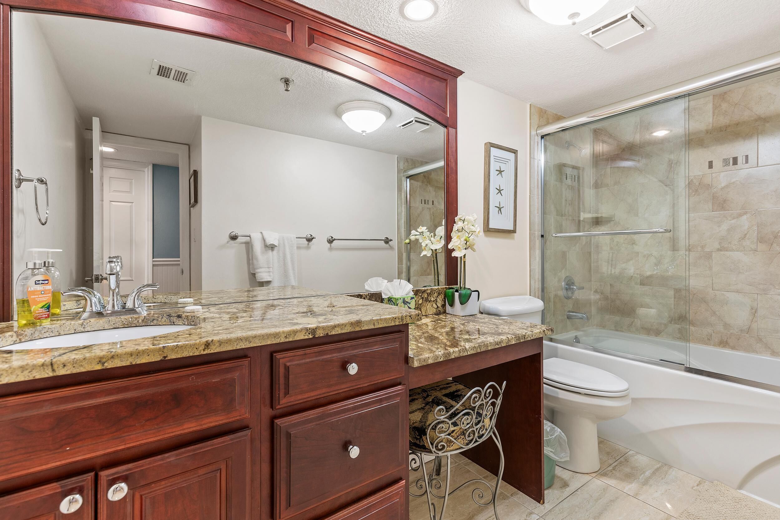 Un baño con encimera de granito, un lavabo blanco, un espejo, cabina de ducha de vidrio y un inodoro.