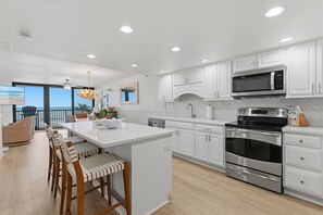 A modern kitchen with white cabinets, a large island, stainless steel appliances, and a view to the outside. - Sunrise 309 (New Smyrna Beach)