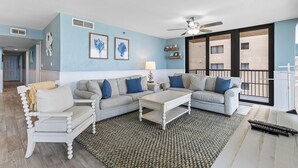 Un salon avec un mur bleu clair, un ventilateur de plafond, une grande fenêtre donnant sur des immeubles, un canapé gris avec des coussins bleus et une table basse blanche.