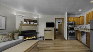 A modern living room with a fireplace, a television, and a kitchen area with wooden cabinets and stainless steel appliances. - 5 Puffins Place: Walk to Beach | Coastal Charm | Dog-Friendly (Cannon Beach)