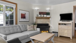 A modern living room with a grey sofa, a coffee table, a fireplace, a television, and a sign that reads "Cappuccino." - 5 Puffins Place: Walk to Beach | Coastal Charm | Dog-Friendly (Cannon Beach)