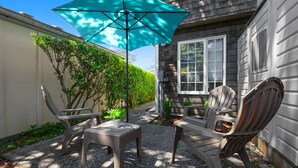 An outdoor seating area with two Adirondack chairs and a table, shaded by a large umbrella, in front of a house with a window and a hedge.